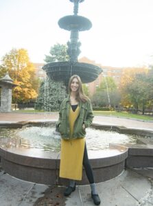 Chelsey Mollin, licensed acupuncturist, herbalist and functional medicine and holistic health practitioner, standing in front of a historic fountain in a city park in Syracuse, New York, during autumn. Wearing a mustard yellow dress layered over black leggings with a green jacket, promoting integrative medicine and natural wellness.